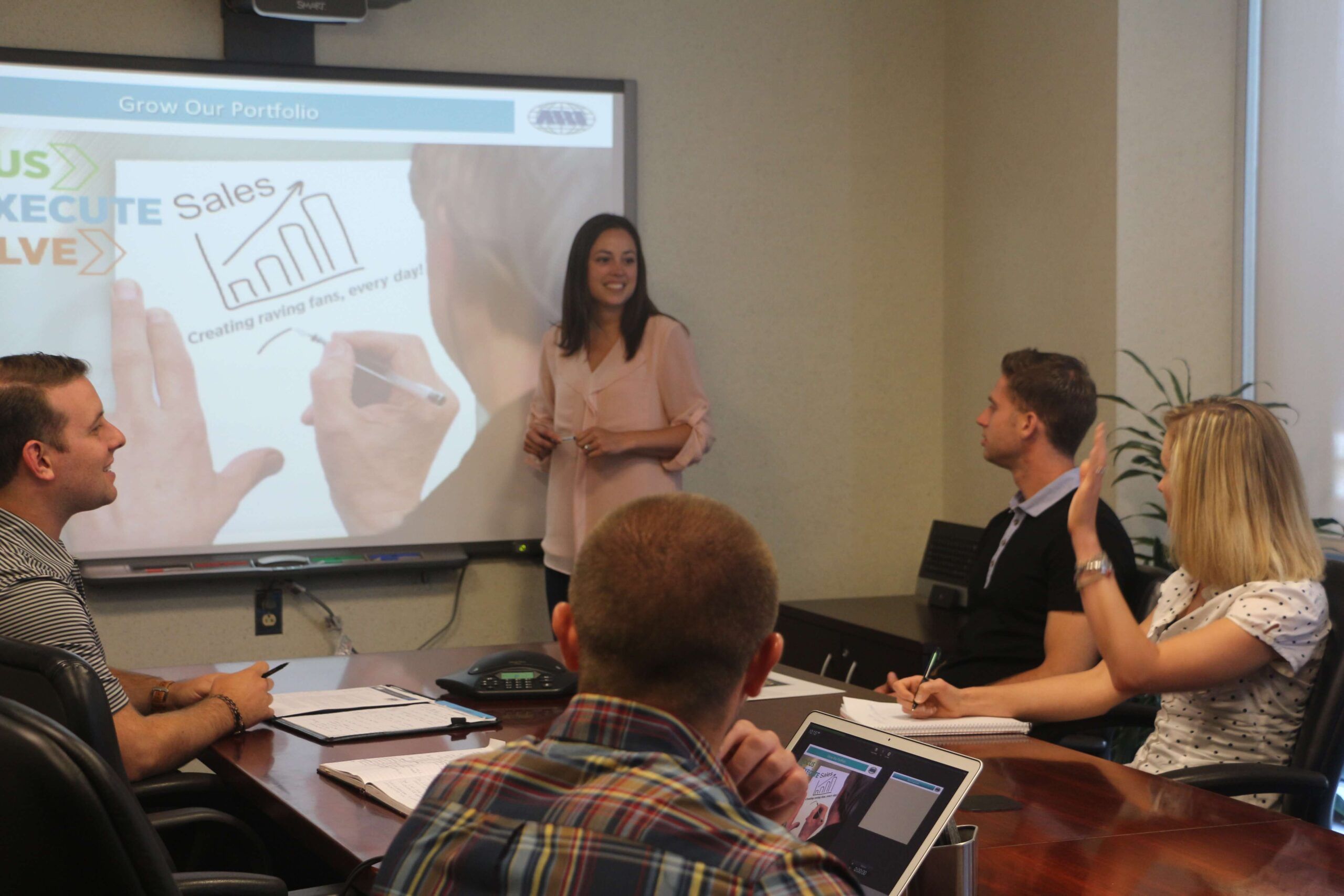 A conference room with viewers sitting around a table watching a presenter utilizing a slide deck.
